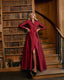A woman stands on wooden stairs in a library wearing a long-sleeved, collared, belted maxi dress in deep burgundy red. The dress has a high leg slit and is styled with black pointed-toe heels.