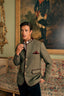 A man wearing a traditional German-style wool blazer in green tweed with dark red trim, standing in an ornate room with a floral chaise lounge and a large historical painting in the background.