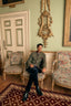A man wearing a green blazer, dark trousers, and black boots sits in a vintage floral armchair in an ornate room with a green wall, gold mirror, and patterned rug.