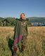 A woman stands in a grassy field wearing a green plaid blazer, olive green scarf, and a large green and purple block-printed shawl, with a scenic bridge and mountains in the background.