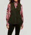 A model wearing an olive green wool vest over a pink and red paisley patterned blouse, paired with dark green trousers, against a light gray studio background.