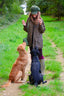 A woman in a brown hunting jacket, green hat, and rubber boots stands on a dirt path in a grassy field, holding treats for two dogs—a golden retriever and a black dog.