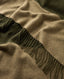 Close-up of a soft woven cape or throw with fringe, shown in warm taupe-beige and dark olive green.