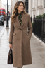 A woman stands on a city sidewalk wearing a long taupe-brown textured overcoat over a dark green floral blouse, holding a dark brown handbag. The background shows a blurred street scene with buildings, a black fence, and a red double-decker bus.