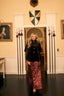 A woman stands in a grand interior hallway wearing a black velvet-like jacket with gold buttons and a long burgundy patterned skirt. The room has cream walls, framed portraits, medieval-style shields, spear shafts, and a dark wood floor.