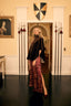 A woman stands in a grand interior hallway wearing a black cape-style top and a long burgundy patterned skirt with a thigh slit. The room has cream walls, decorative spear shafts, a shield, framed artwork, and a dark floor.