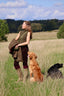 A woman standing in a grassy field with two dogs, wearing an olive green sleeveless top, dark brown pants, and tall green boots, while holding a brown jacket. Trees and a blue sky are visible in the background.