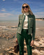 Woman in a green jacket and pants standing on a rocky beach with a blue sky.