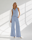 A woman models a light blue sleeveless vest and matching wide-leg trousers set, standing in a bright white studio with diagonal window light shadows.
