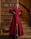 A woman stands in a grand library wearing a long, deep burgundy satin-like wrap dress. She is posed next to a tall bookshelves and a wooden staircase behind her.