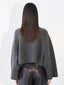 A woman viewed from the back wearing a dark gray textured knit cropped sweater and shiny black pants against a plain white background.