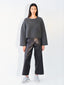 A model stands against a plain white studio background wearing a dark gray knit cropped sweater, black wide-leg leather pants, and white sneakers.