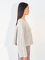 A woman shown in side profile wearing a light beige textured knit sweater with wide draped sleeves and gray trousers, standing against a plain white wall.