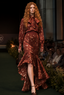 A model walks a fashion runway wearing a rust-brown patterned long-sleeve blouse and matching high-low ruffled skirt set, with a bow at the collar. The outfit has a subtle metallic sheen and is paired with dark brown pointed-toe heels.