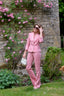 Sophie Stanbury in a pink suit standing in a garden with pink flowers and a stone wall.