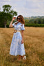 A woman wearing a light blue and white floral midi dress with a tan belt stands in a dry grassy field under a cloudy sky.