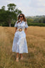A woman wearing a light blue and white patterned midi shirt dress with a wide tan belt stands in a dry grassy field with trees in the background.