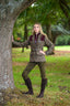 A woman posing outdoors in a park next to a large tree, wearing a brown tweed blazer with dark burgundy shoulder panels, olive green trousers, and dark brown knee-high boots.