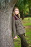 A woman posing outdoors against a large tree trunk, wearing a brown and beige plaid tweed blazer with dark burgundy shoulder panels, olive green trousers, and a dark top.