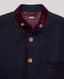 Close-up of a navy blue jacket with deep burgundy velvet trim on the collar and pocket, accented by gold-tone buttons and a gold-tone chain detail inside the collar.