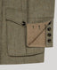Close-up of a herringbone tweed jacket in olive-brown and beige tones, showing a front flap pocket with a dark button and a smaller angled pocket with visible stitching and dark buttons.