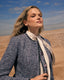Woman in a boucle coat and blouse standing in a beach landscape with a blue sky.