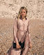 Woman in a long, light pink dress standing on a pebbly beach.