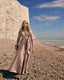 Woman in a long, light pink dress standing on a pebbly beach.