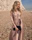 Woman in a long, light pink dress standing on a pebbly beach.