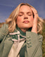 Woman in a green blazer with a scarf, standing outdoors against a blue sky.