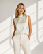 A woman modeling a sleeveless high-neck satin blouse in a warm off-white/cream color, paired with matching cream trousers, set against a bright white wall with soft window shadow patterns.