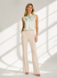 A woman stands in a bright studio wearing a sleeveless pale mint satin blouse with a large bow at the collar and light beige wide-leg trousers. Sunlight casts diagonal shadows across the white floor and wall.