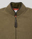 Close-up of a cape zip-front jacket with a high stand collar, made from textured olive-brown fabric, featuring a dark brown leather tassel zipper pull and a dark olive-green inner collar lining.