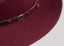 Close-up of A dark burgundy felt fedora hat with a thin dark brown band and a small gold buckle, shown against a solid light gray background.