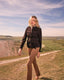 Woman in a brown jacket and beige pants standing on a grassy hill with a scenic landscape in the background.