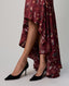 A close-up of a model's legs wearing a high-low skirt in deep burgundy with a botanical leaf pattern, paired with black pointed-toe stiletto pumps against a light gray studio background.