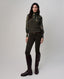 A woman modeling a dark olive green ribbed knit sweater vest over a long-sleeve floral shirt, paired with dark olive green riding pants and dark brown knee-high boots, against a light gray studio background.