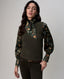 A woman modeling a dark olive green ribbed knit vest over a long-sleeve button-up shirt with a green camouflage print and a matching green neck scarf, paired with dark brown pants.