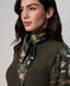 A woman wearing a dark green knit sweater vest over a long-sleeve blouse with a feather print, accessorized with a matching patterned neck scarf, against a neutral gray background.