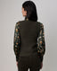 A woman shown from the back wearing a dark olive green ribbed knit sweater vest over a long-sleeve blouse with a brown and beige feather print, paired with dark brown pants, against a light gray studio background.