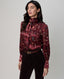 A woman modeling a long-sleeve burgundy satin blouse with a tie-neck collar and an allover feather print, paired with dark brown trousers and a brown belt, against a neutral gray studio background.