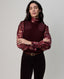 A woman modeling a burgundy mock-neck top with sheer patterned long sleeves and a ribbed knit vest, paired with dark pants and a brown belt, against a plain light gray background.