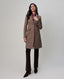 A woman modeling a mid-length taupe-brown wool coat over a patterned blouse and dark brown trousers, standing against a light gray studio background.