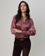 A woman modeling a long-sleeve button-up shirt with a metallic pink and purple paisley pattern, paired with dark brown trousers and a brown belt, against a solid light gray background.