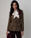 A woman models a brown tweed belted blazer with a white bow tie and dark burgundy trousers against a neutral gray background.