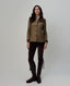 A woman modeling a brown and beige plaid jacket with dark brown shoulder panels, paired with dark brown pants and tall dark brown riding boots, against a light gray studio background.