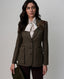 A woman models a brown tweed blazer with a three-button front, flap pockets, and a decorative floral lapel pin, paired with a cream satin bow tie and dark trousers against a light gray studio background.