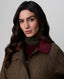 A woman with long, wavy dark brown hair is shown from the chest up, wearing a brown textured wool-blend jacket with a ribbed burgundy corduroy collar over a dark olive green turtleneck sweater, set against a plain light gray studio background.