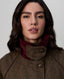 A close-up portrait of a woman wearing a brown tweed jacket with a high ribbed burgundy corduroy collar over a dark green turtleneck, set against a plain light gray background.