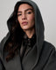 A close-up portrait of a woman wearing a dark charcoal gray oversized hooded coat over a black satin bow-neck blouse, set against a plain light gray studio background.